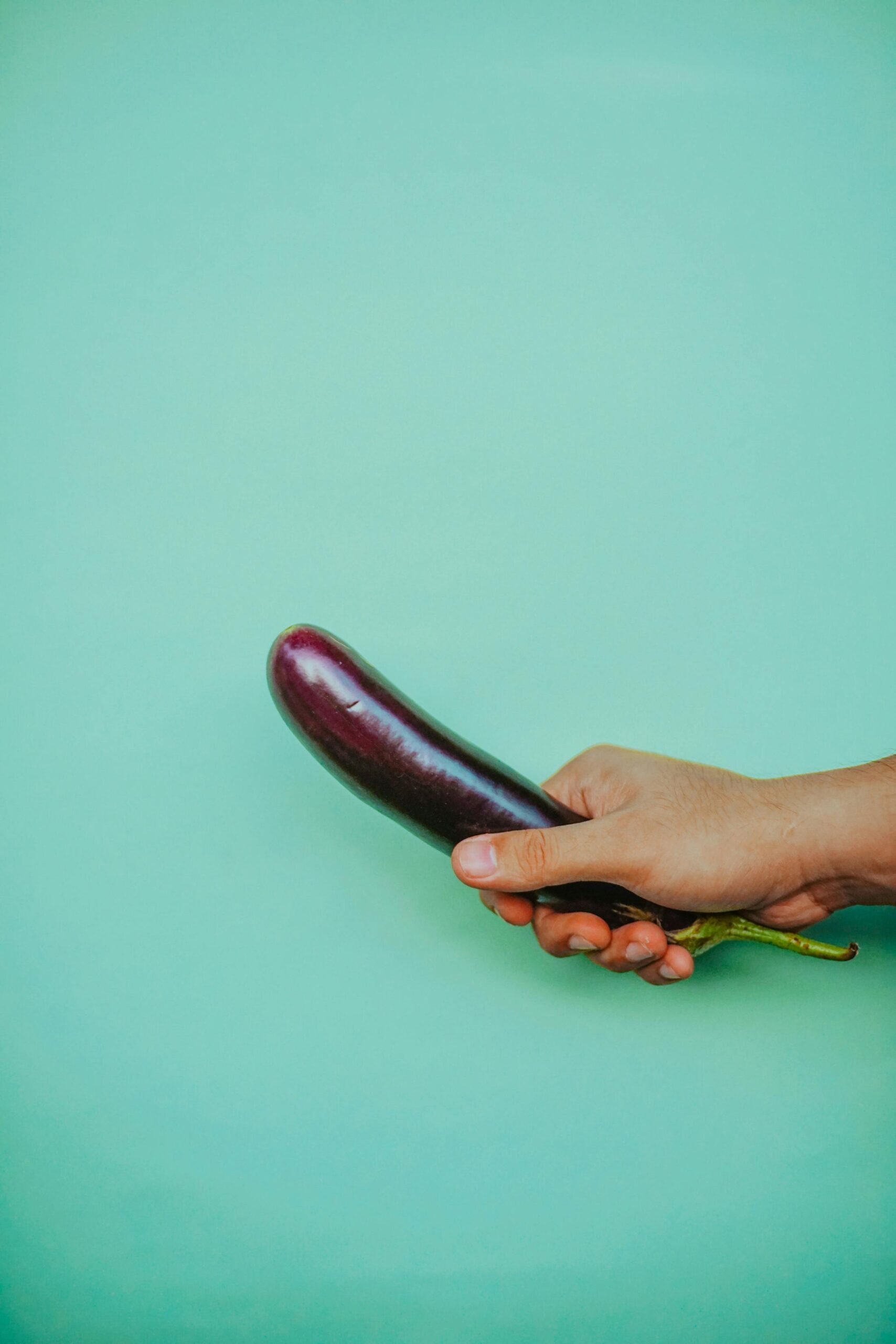 Close-up of a hand holding an eggplant against a solid green background.